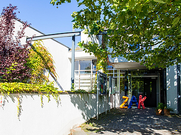 Eingangsbereich des Fröbel‑Kindergartens & Familienzentrums ZAK in Bergisch Gladbach. Vor dem Eingang stehen bunte, große Buchstaben, die „ZAK“ bilden. Bäume und Pflanzen werfen Schatten auf den Weg.